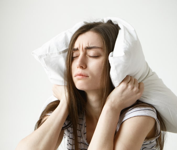 pretty-student-woman-in-striped-t-shirt-trying-to-have-some-sleep-sitting-in-bedroom-with-pillow-on-her-head-closing-eyes